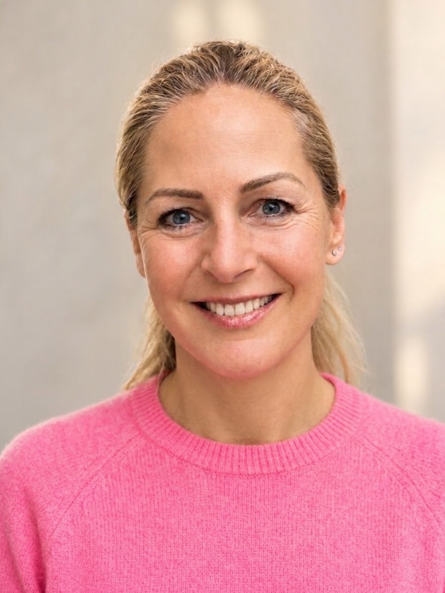 Anne Brenner lächelt offen in die Kamera, im pinkfarbenen Pullover vor heller Wand